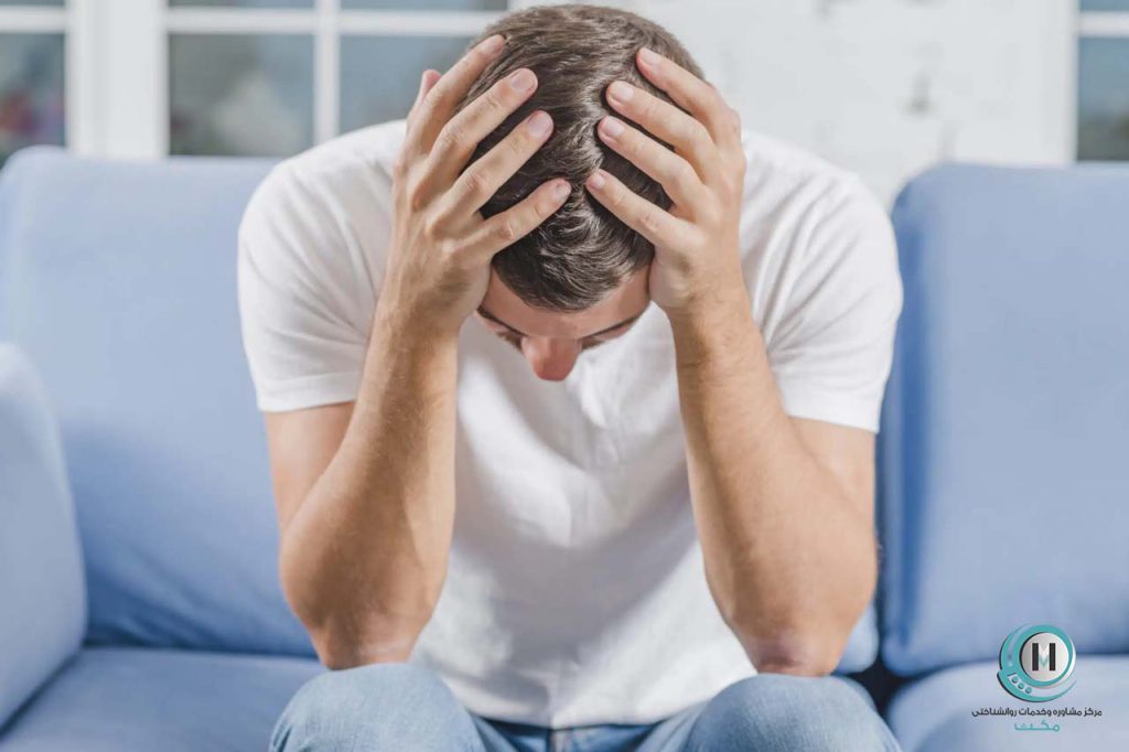 مردی با لباس سفید که اضطراب دارد و سرش را بین دستانش گرفته و روی مبل نشسته است